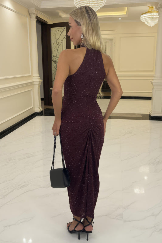Woman in a burgundy one-shoulder dress holding a black handbag in an elegant interior setting.