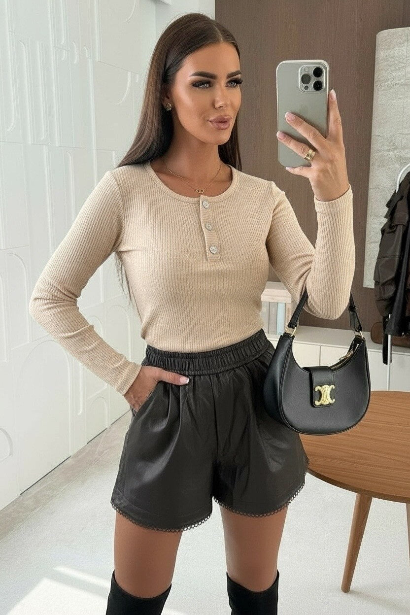 Woman taking a mirror selfie wearing a beige long-sleeve top and black shorts, holding a black handbag.