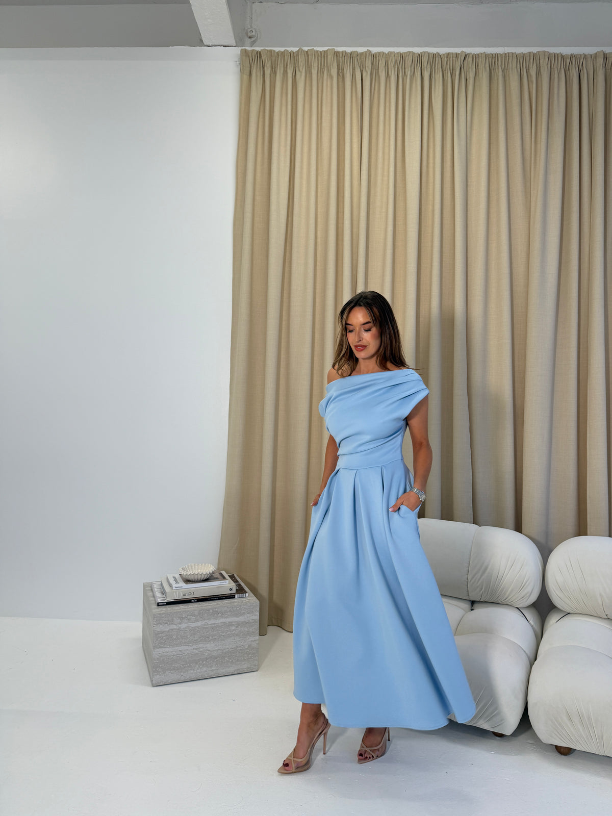 Woman in a light blue dress standing in a room with beige curtains and a white sofa.
