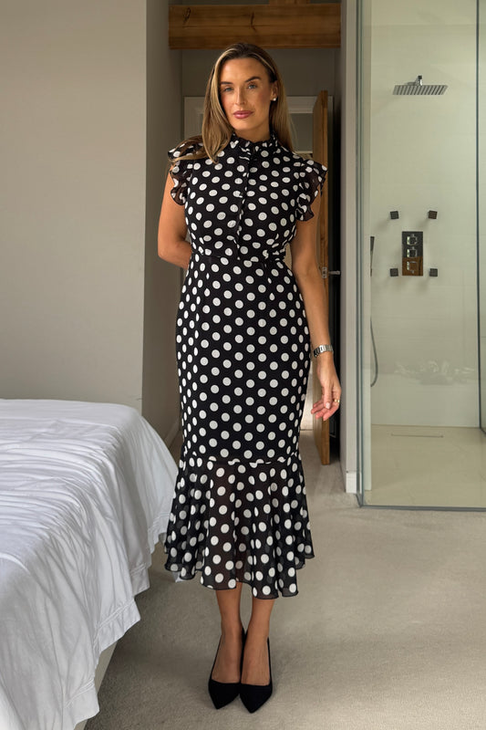 Woman wearing a black and white polka dot dress in a bedroom setting.