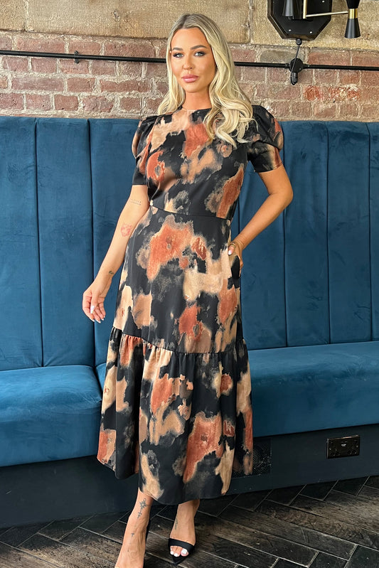 Woman wearing a patterned dress standing in front of a blue couch with a brick wall background.