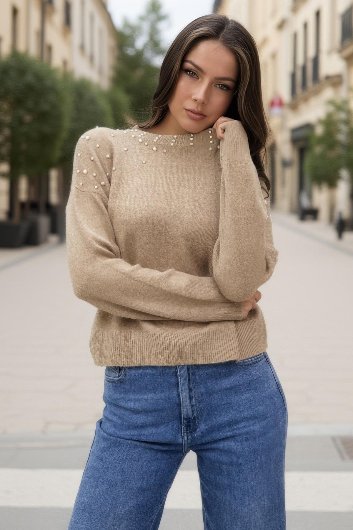 Woman wearing a beige sweater with pearl embellishments and blue jeans on a city street.