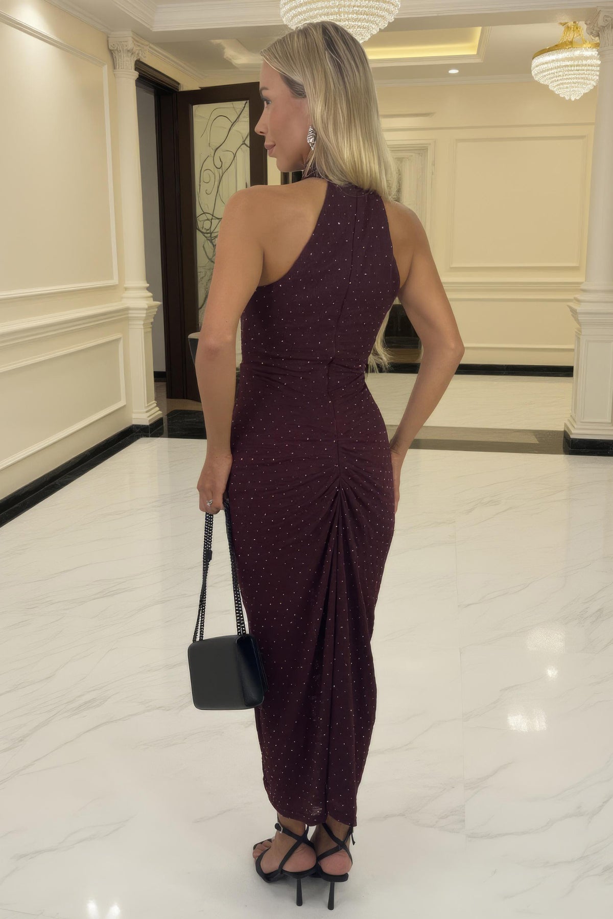 Woman in a burgundy one-shoulder dress holding a black handbag in an elegant interior setting.
