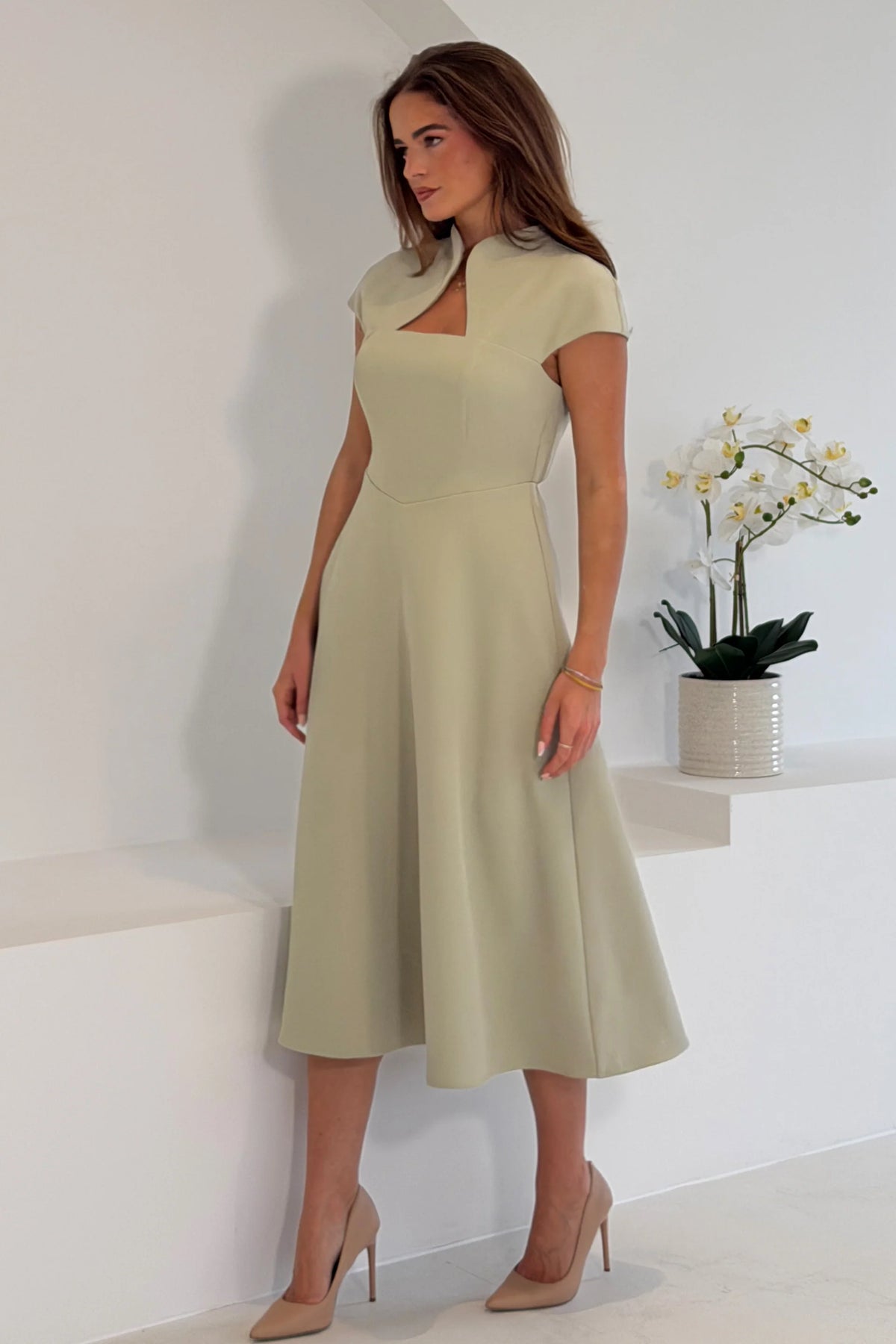 Woman wearing a light green dress standing in a minimalistic room with a plant.