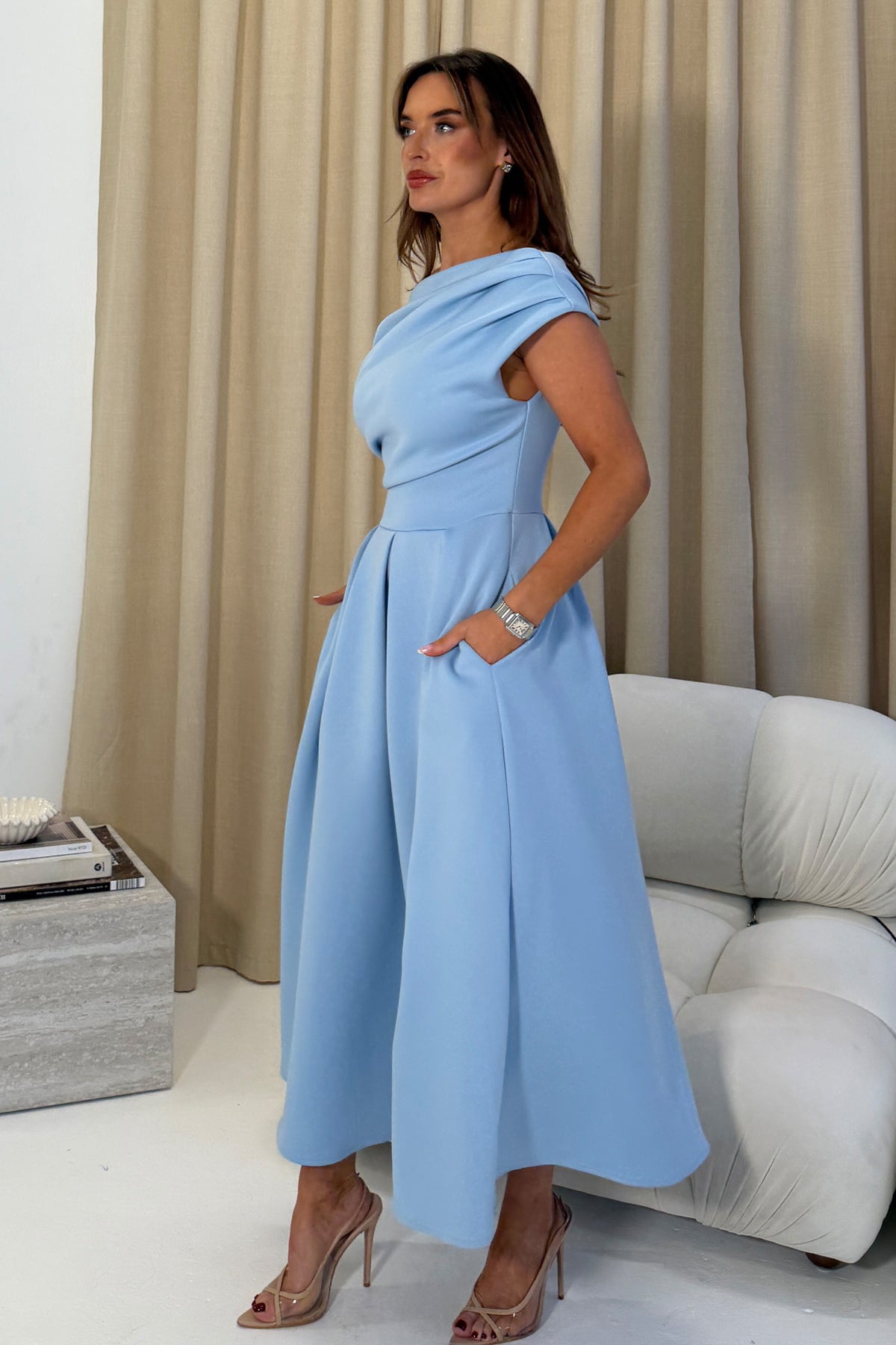 Woman wearing a light blue dress standing in a room with beige curtains and a white sofa.