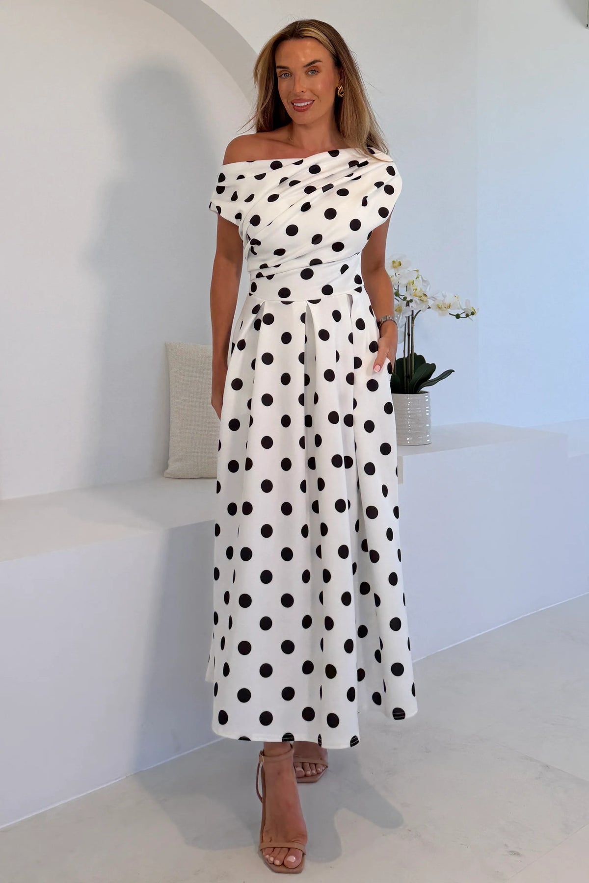 Woman wearing a white off-shoulder dress with black polka dots in a bright interior setting.