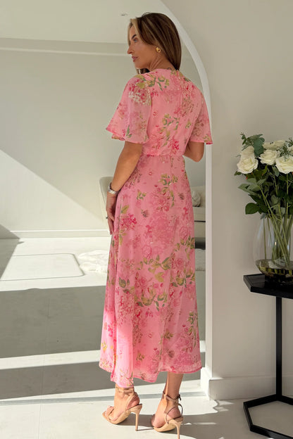 Woman in a pink floral dress standing in a sunlit room with a vase of flowers on a table.
