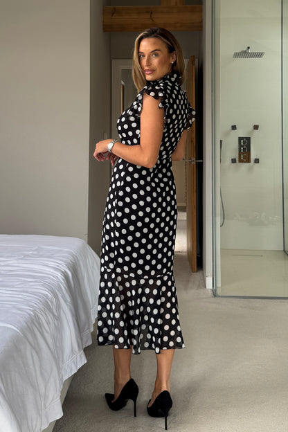 Woman in a black and white polka dot dress standing in a bedroom.
