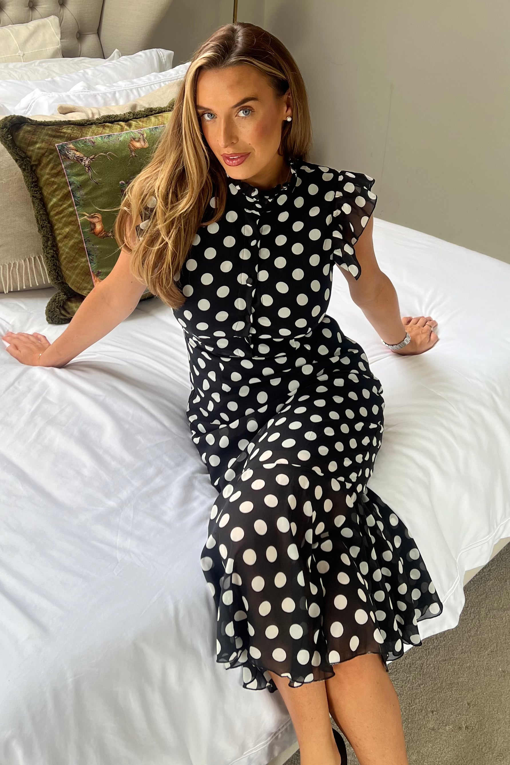 Woman in a black polka dot dress sitting on a bed.