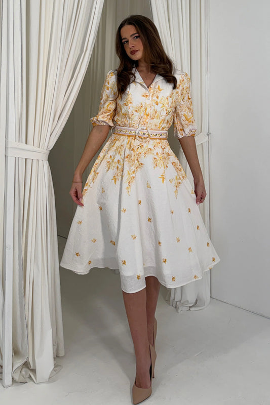 Woman wearing a white dress with yellow floral patterns in a room with white curtains.