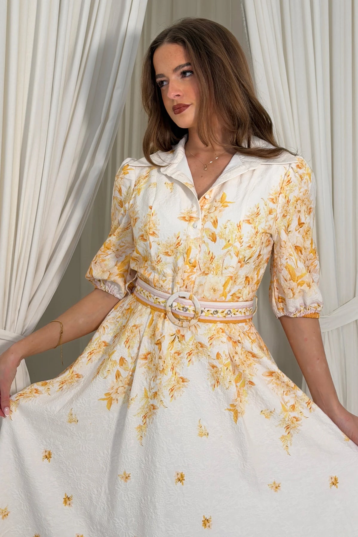 Woman wearing a white dress with yellow floral pattern in a softly lit room.