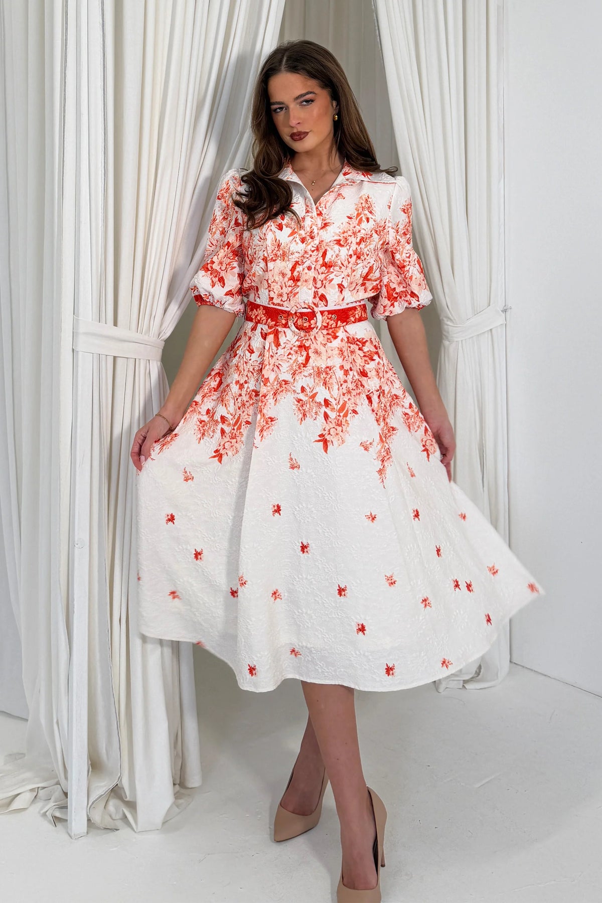 Woman wearing a white dress with red floral patterns in a room with white curtains.