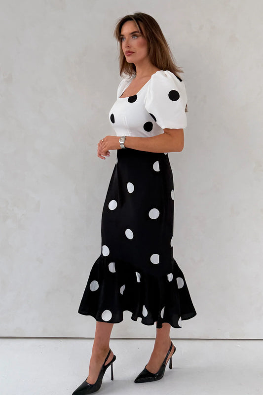 Woman wearing a white top with black polka dots and a black skirt with white polka dots against a light gray background.