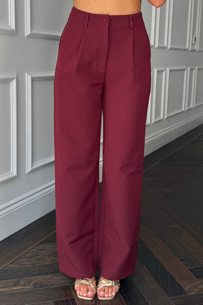 Maroon high-waisted pants worn by a person standing in a room with gray paneled walls.