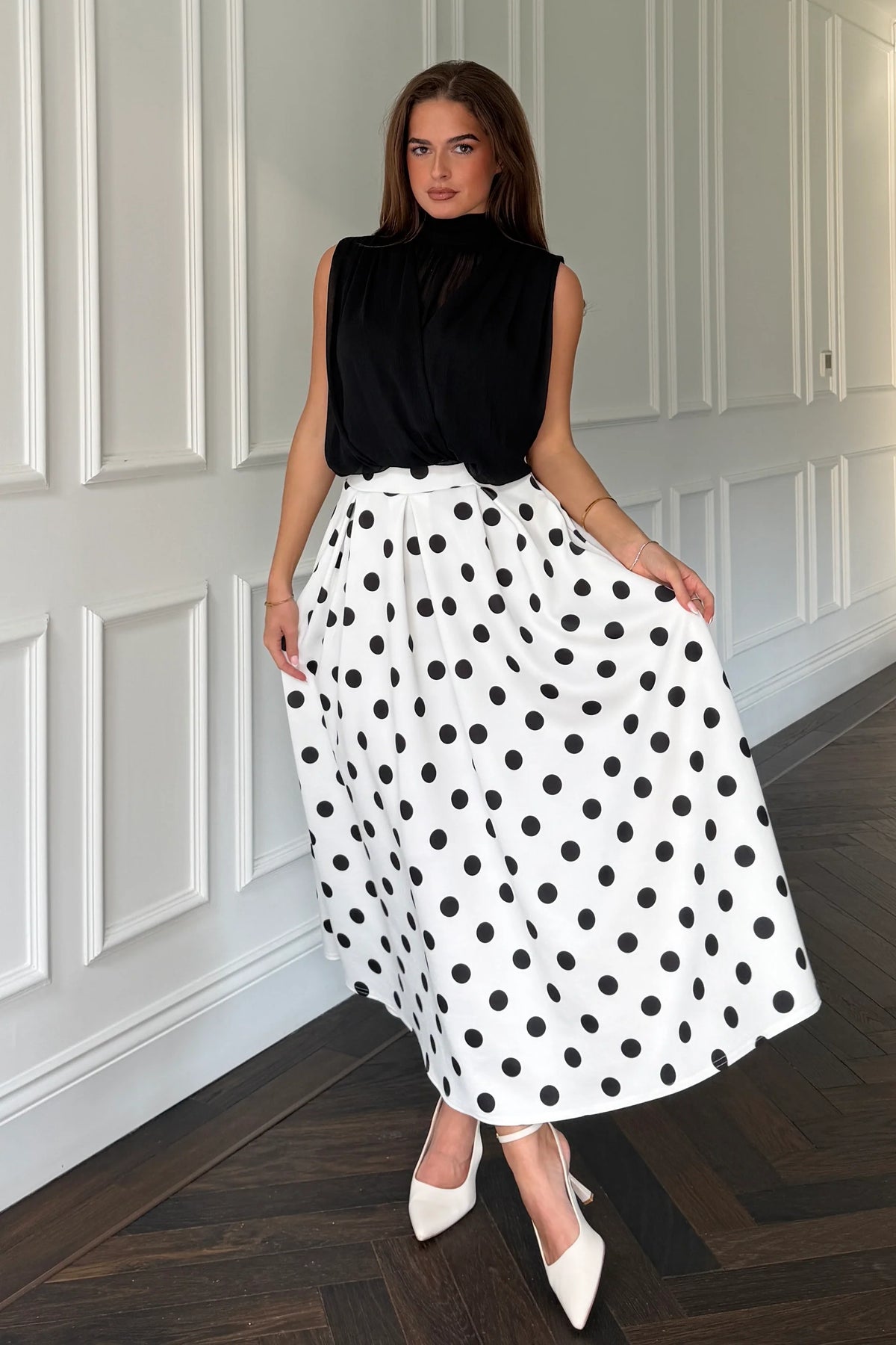 Woman wearing a black sleeveless top and white polka dot skirt in a room with white paneled walls.