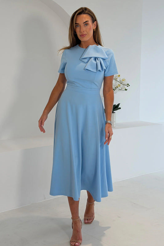 Woman wearing a light blue dress with a large bow detail in a minimalistic setting.