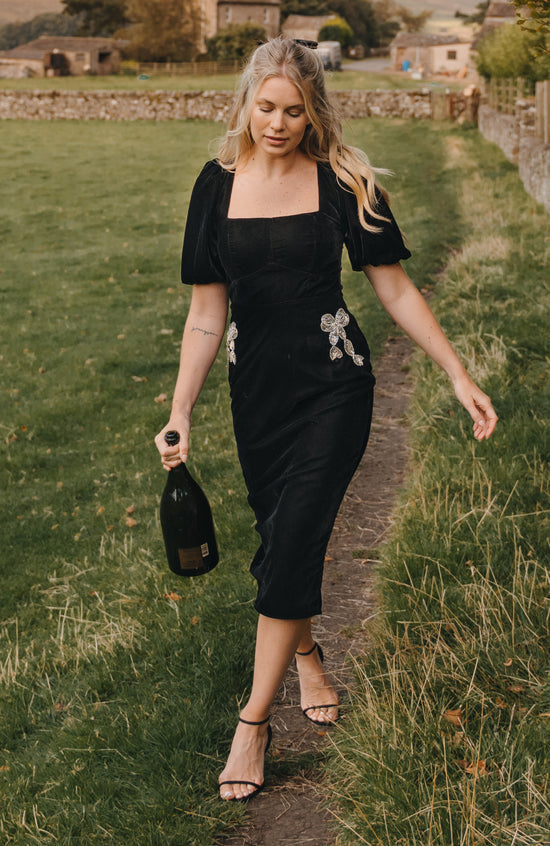 Woman in a black dress walking along a path in a rural setting, holding a bottle.
