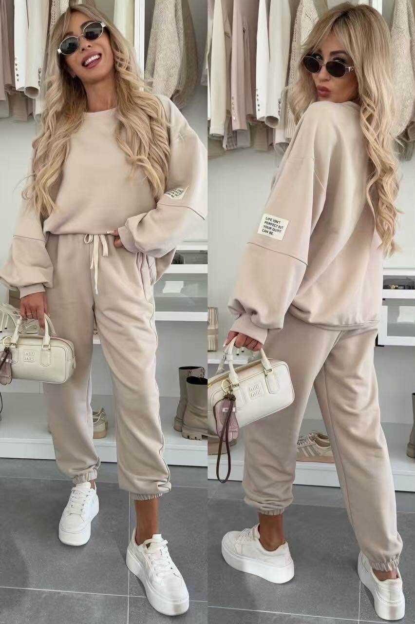 Woman wearing a beige tracksuit with sunglasses and a handbag, standing in front of a rack of clothes.