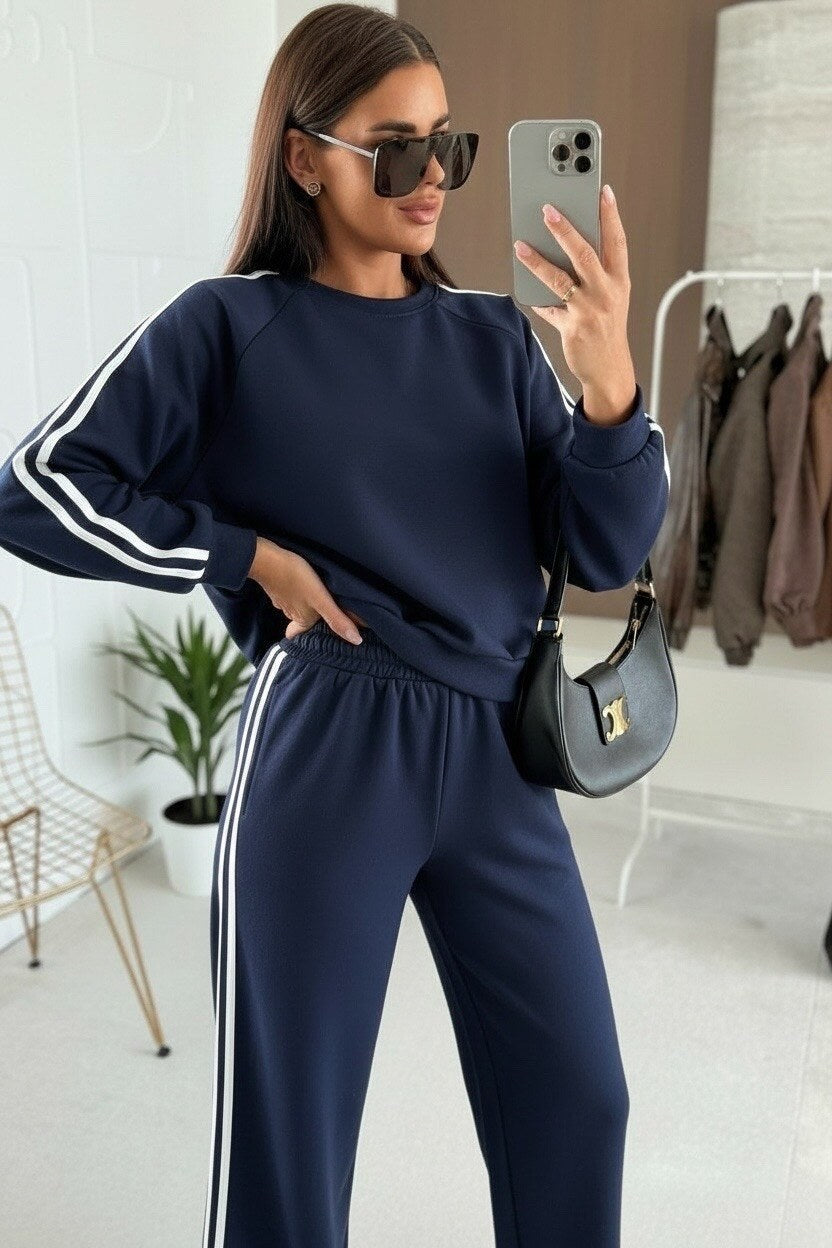Woman in navy tracksuit taking a mirror selfie in a room with a plant and coat rack.