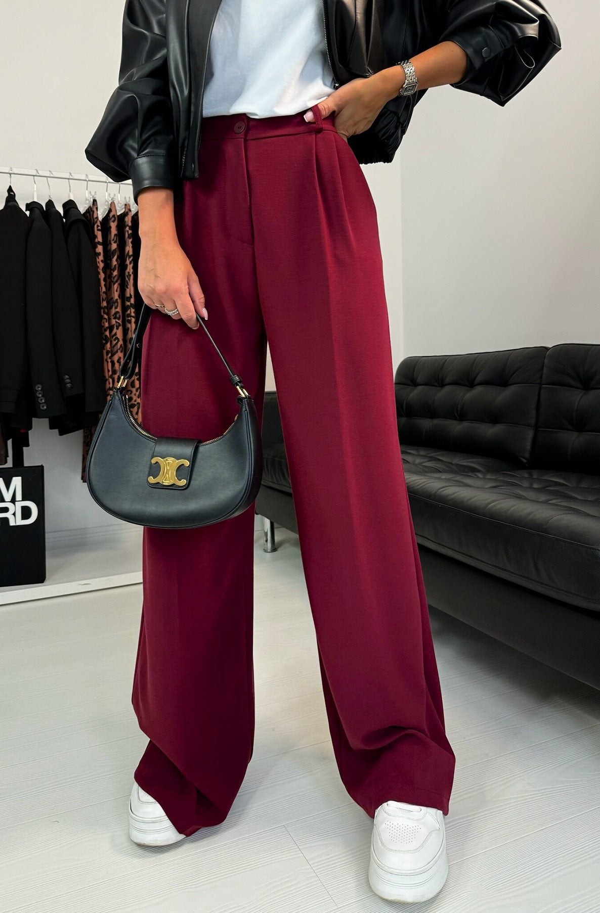 Person wearing burgundy pants with a black jacket and white sneakers, holding a black handbag.