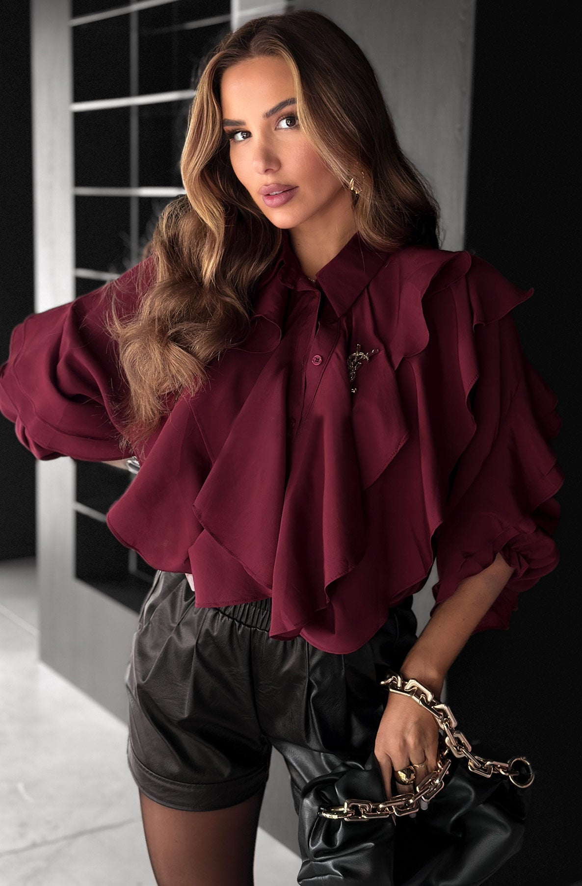 Woman wearing a burgundy blouse with ruffled details and black shorts, holding a black handbag.